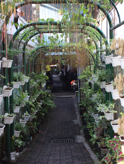 A narrow pathway within a green, arch-covered garden corridor features a brick-paved walkway flanked by lush potted plants and shrubs on both sides. The arch structure is covered with hanging and clim