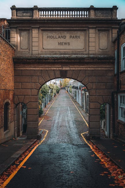 A narrow cobblestone street viewed through an archway of a historic brick building with a carved stone inscription reading 'HOLLAND PARK MEWS'. The street is lined with front gardens enclosed by low f