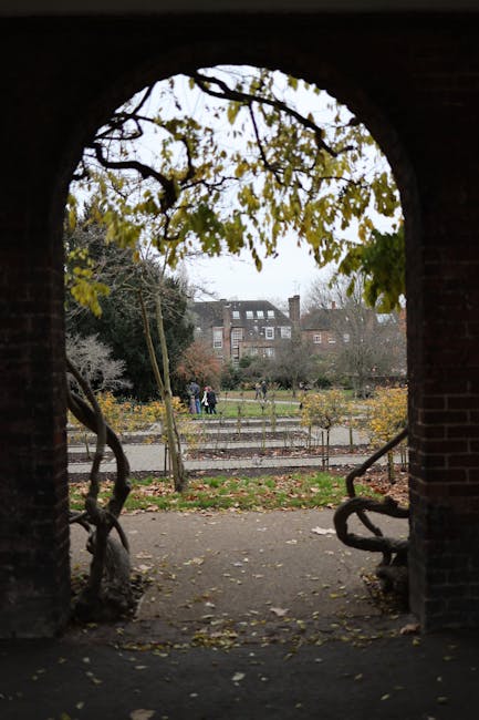 A narrow cobblestone street viewed through an archway of a historic brick building with a carved stone inscription reading 'HOLLAND PARK MEWS'. The street is lined with front gardens enclosed by low f
